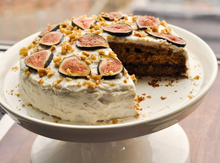 Fig cake on a white pedestal cake platter frosted with white icing with sliced figs on top.