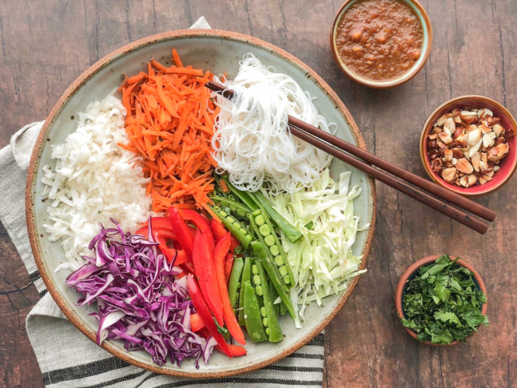 Serving bowl filled with shredded carrots, vermicelli noodles, sliced red cabbage, sliced red peppers, and pea pods that are halved lengthwise, all arranged in their own part of the bowl. Chopsticks sit on top and three small bowls are off to the side filled with cilantro, peanuts, and dressing.