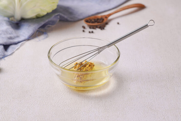 A whisk sits on top of a clear bowl containing olive oil and wholegrain mustard.