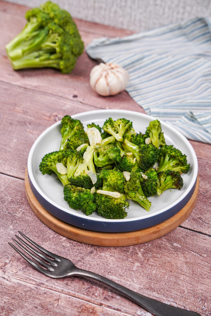 A plate of roasted broccoli with olive oil, sliced garlic and salt and pepper, surrounded by garlic cloves, a raw broccoli floret and a fork.