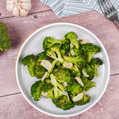 A large plate of roasted broccoli with olive oil, sliced garlic and salt and pepper, surrounded by garlic cloves and a broccoli floret.