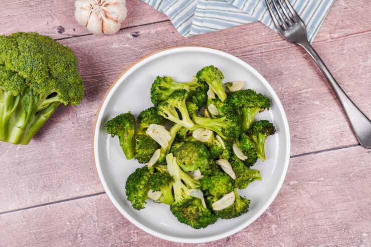 A large plate of roasted broccoli with olive oil, sliced garlic and salt and pepper, surrounded by garlic cloves and a broccoli floret.