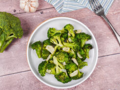 A large plate of roasted broccoli with olive oil, sliced garlic and salt and pepper, surrounded by garlic cloves and a broccoli floret.