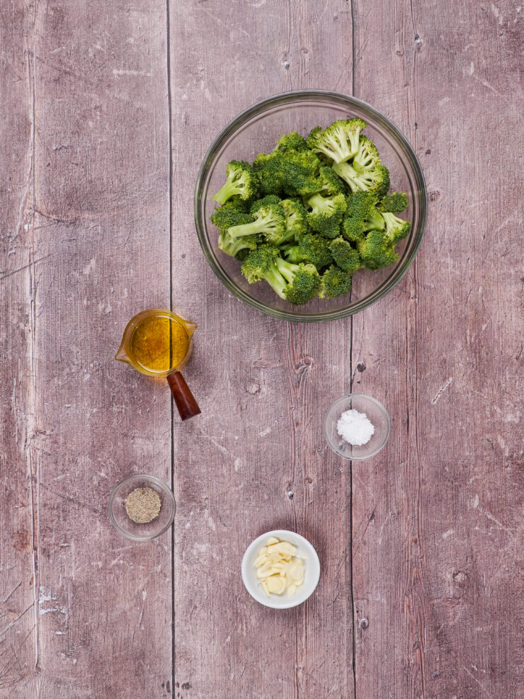 Ingredients for roasted broccoli, including a cup of olive oil, a bowl of raw broccoli, peeled garlic cloves, salt and pepper.