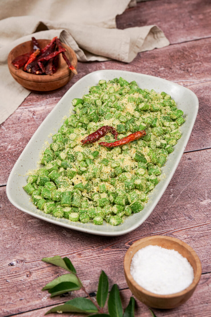 Rectangular white plate filled with cooked sliced string beans and shredded coconut with a garnish of two dried red chilis. A wooden bowl filled with shredded coconut sits in front while a wooden bowl of dried red chilis sits in behind.