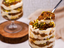Two tall dessert glasses of carrot halwa trifle with a spoon dipping into one of them. You can see the layers of the carrot mixture and cream mixture in the glasses.