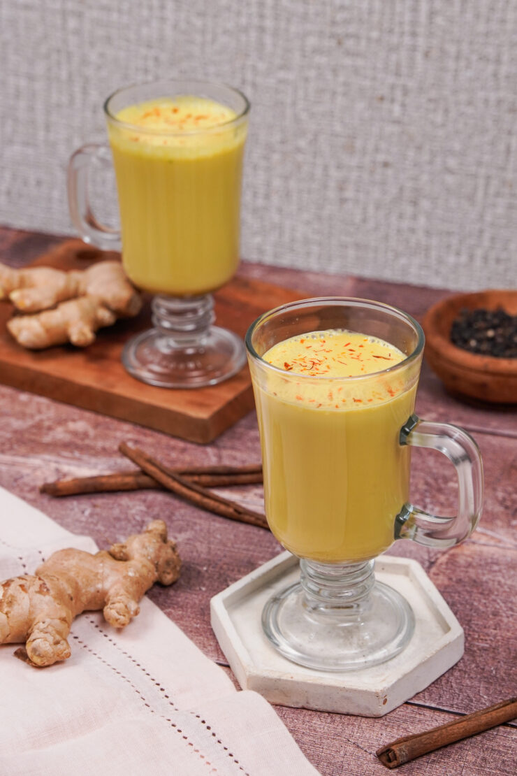 Two clear glass mugs with handles filled with golden yellow vegan turmeric latte with a couple ginger roots and cinnamon sticks around them.