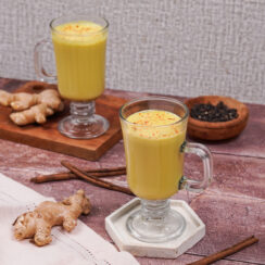 Two handled glasses filled with golden vegan turmeric latte with ginger root and cinnamon sticks surrounding them.