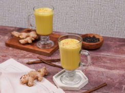 Two handled glasses filled with golden vegan turmeric latte with ginger root and cinnamon sticks surrounding them.