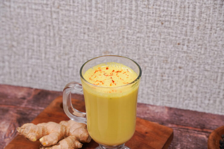 Closeup view of a clear glass mug filled with golden vegan turmeric latte sitting on a wooden board with ginger root sitting on the side.
