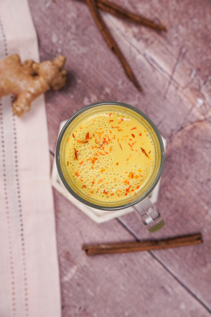 Overhead shot of a clear glass mug filled with golden colored vegan turmeric latte with a ginger root and cinnamon sticks strewn around it.