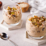 Two clear glass dishes are filled with creamyvegan kheer pudding, topped with chopped nuts. The smooth texture of the pudding is visible through the sides of the glass, and a decorative spoon rests on the saucer next to the dish.