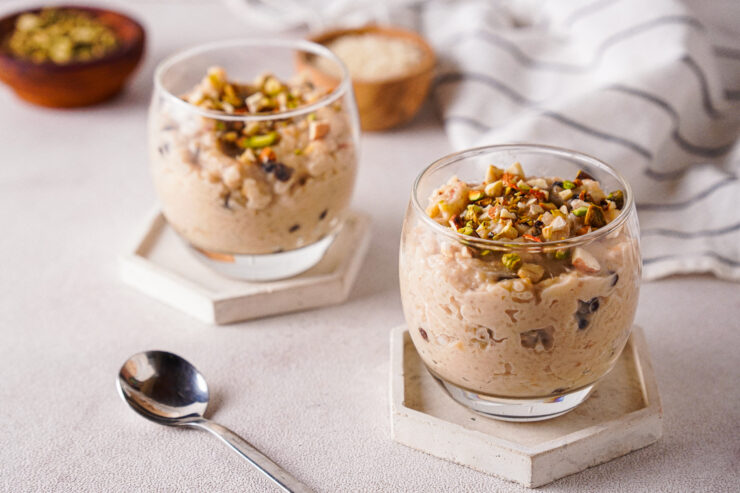 Two clear glass dishes are filled with creamy vegan kheer pudding, topped with chopped nuts. The smooth texture of the pudding is visible through the sides of the glass, and a decorative spoon rests on the saucer next to the dish.