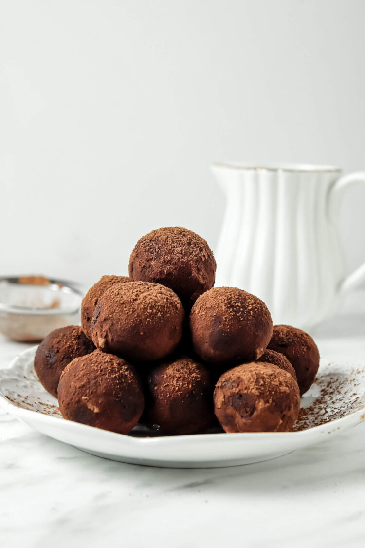 Pile of cocoa dusted coconut chocolate truffles on a round white plate with a white pitcher behind.