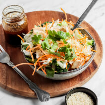 Bowl of Asian cabbage salad filled with shredded cabbage and carrots, and cilantro leaves on top. An open jar of dressing sits behind the bowl.