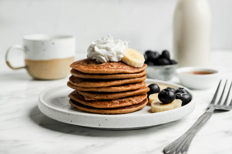 Stack of banana pancakes on a white plate with blueberries, sliced banana, and whipped coconut cream
