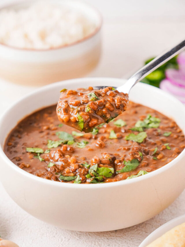 White bowl of dal makhani garnished with chopped cilantro. A spoon hovers above the bowl and is full of the spiced lentils while a bowl of rice sits in the background.