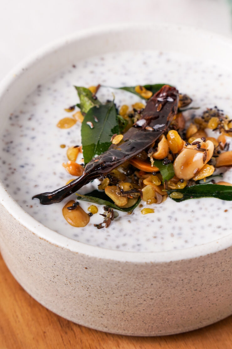 White bowl filled with Greek yogurt garnished with peanuts, mustard seeds, curry leaves, red chili, and ginger