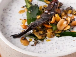 White bowl filled with Greek yogurt garnished with peanuts, mustard seeds, curry leaves, red chili, and ginger
