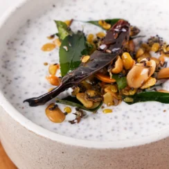 White bowl filled with Greek yogurt garnished with peanuts, mustard seeds, curry leaves, red chili, and ginger