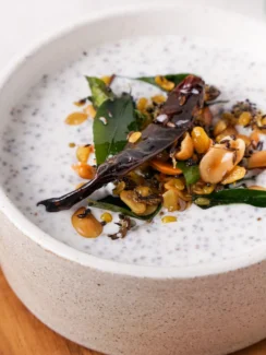 White bowl filled with Greek yogurt garnished with peanuts, mustard seeds, curry leaves, red chili, and ginger