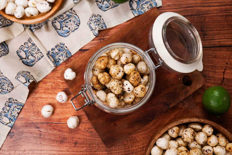 Overhead shot of a jar filled with masala makhana on a wooden board with a bowl of makhana and whole lime in front of it
