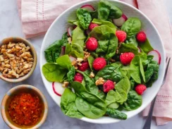 Spinach salad in a white bowl with raspberries, radishes, and nuts on top, and bowls of nuts and raspberry puree on the side
