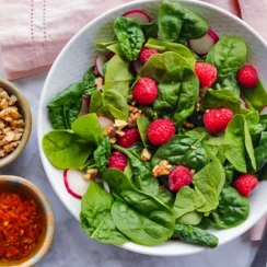 Spinach salad in a white bowl with raspberries, radishes, and nuts on top, and bowls of nuts and raspberry puree on the side