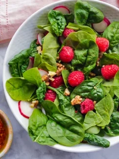 Spinach salad in a white bowl with raspberries, radishes, and nuts on top, and bowls of nuts and raspberry puree on the side