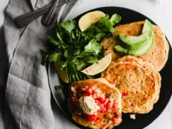 Three chickpea pancakes topped with cilantro, avocado and salsa are served on a black plate.