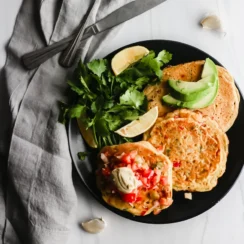 Three chickpea pancakes topped with cilantro, avocado and salsa are served on a black plate.