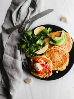Three chickpea pancakes topped with cilantro, avocado and salsa are served on a black plate.