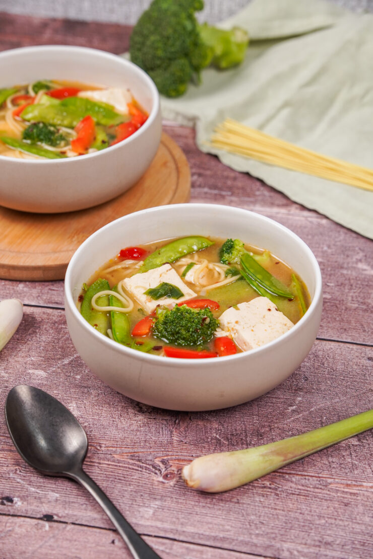 Two bowls of miso soup with tofu, surrounded by ingredients like broccoli and green onion.