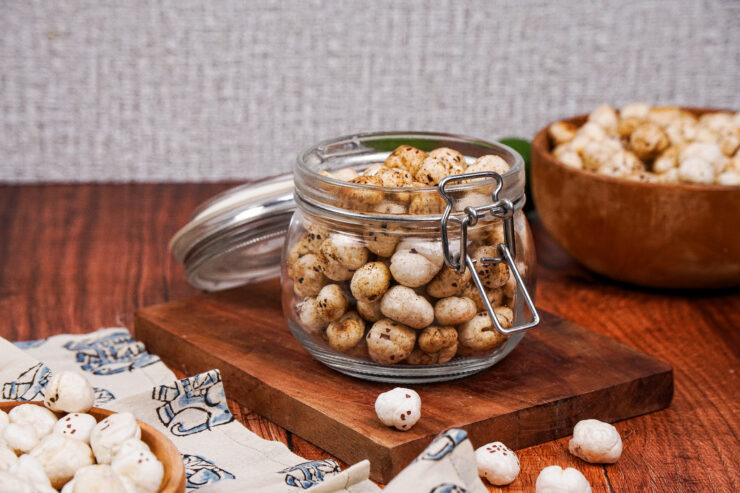 Jar of spiced masala makhana on top of a wooden board with a wooden bowl of makhana behind it