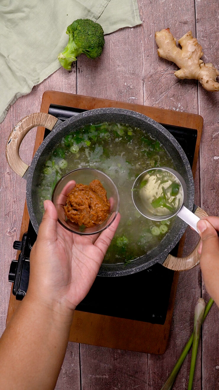Hands add miso paste and green onions to a miso soup simmering on a stovetop.
