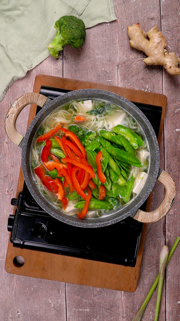 Chopped red peppers and snow peas have recently been added to a miso soup simmering on a stovetop.