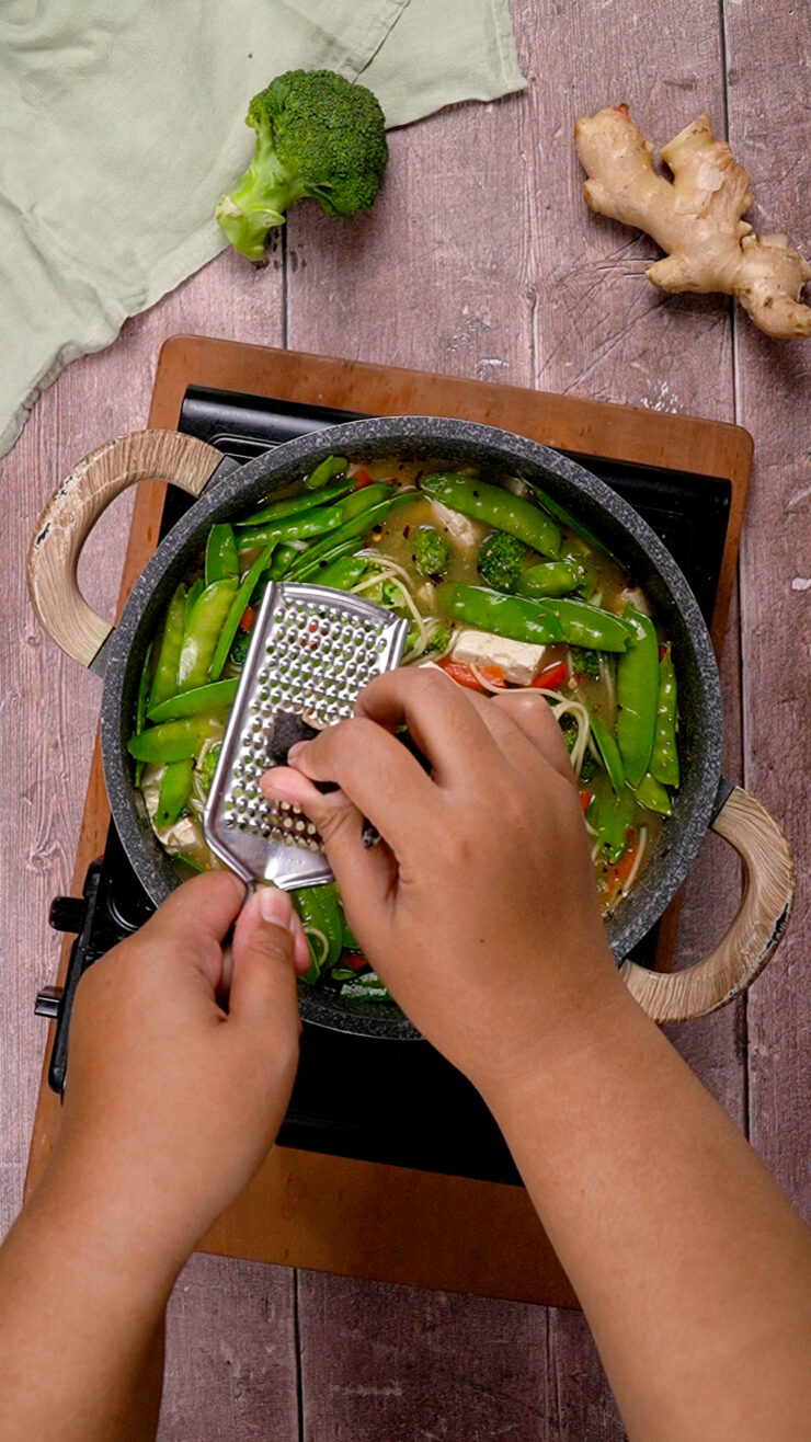 Hands grate dried mushroom over miso soup simmering on a stovetop.