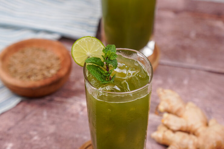 A tall glass of green hued jaljeers with a lime slice on the side of the glass and a mint sprig sitting on top. Another glass sits in behind while a gingerroot is sitting on the table off to the side.