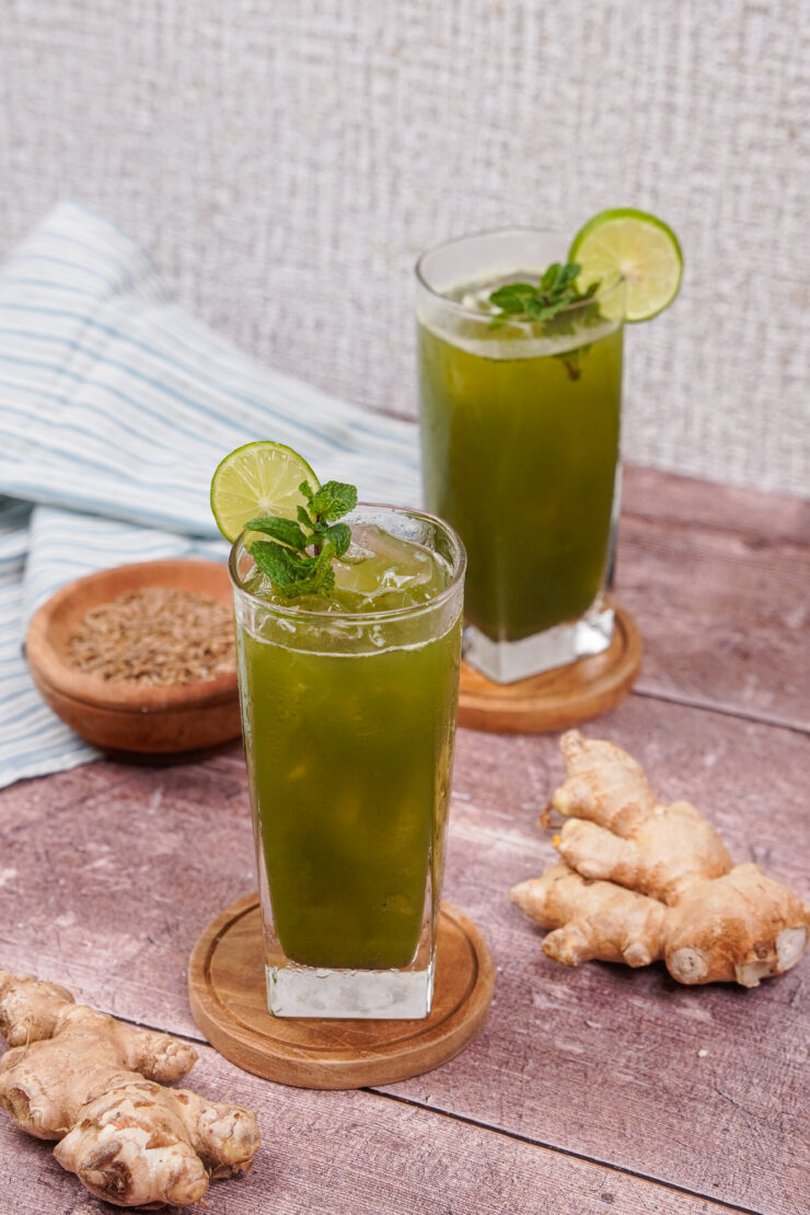 Two tall glasses filled with jaljeera water with lemon slices on the glasses and mint sprigs sitting on top. A whole gingerroot sits on the side.