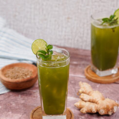 Two tall glasses filled with jaljeera with lime slices on the rims and sprigs of mint in the glasses. A ginger root sits between them.