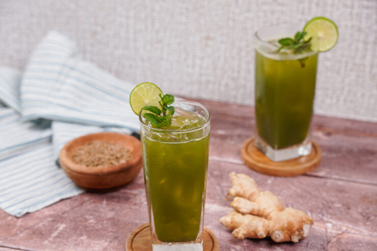 Two tall glasses filled with jaljeera with lime slices on the rims and sprigs of mint in the glasses. A ginger root sits between them.