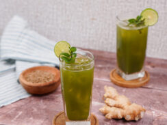 Two tall glasses filled with jaljeera with lime slices on the rims and sprigs of mint in the glasses. A ginger root sits between them.