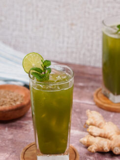 Two tall glasses filled with jaljeera with lime slices on the rims and sprigs of mint in the glasses. A ginger root sits between them.