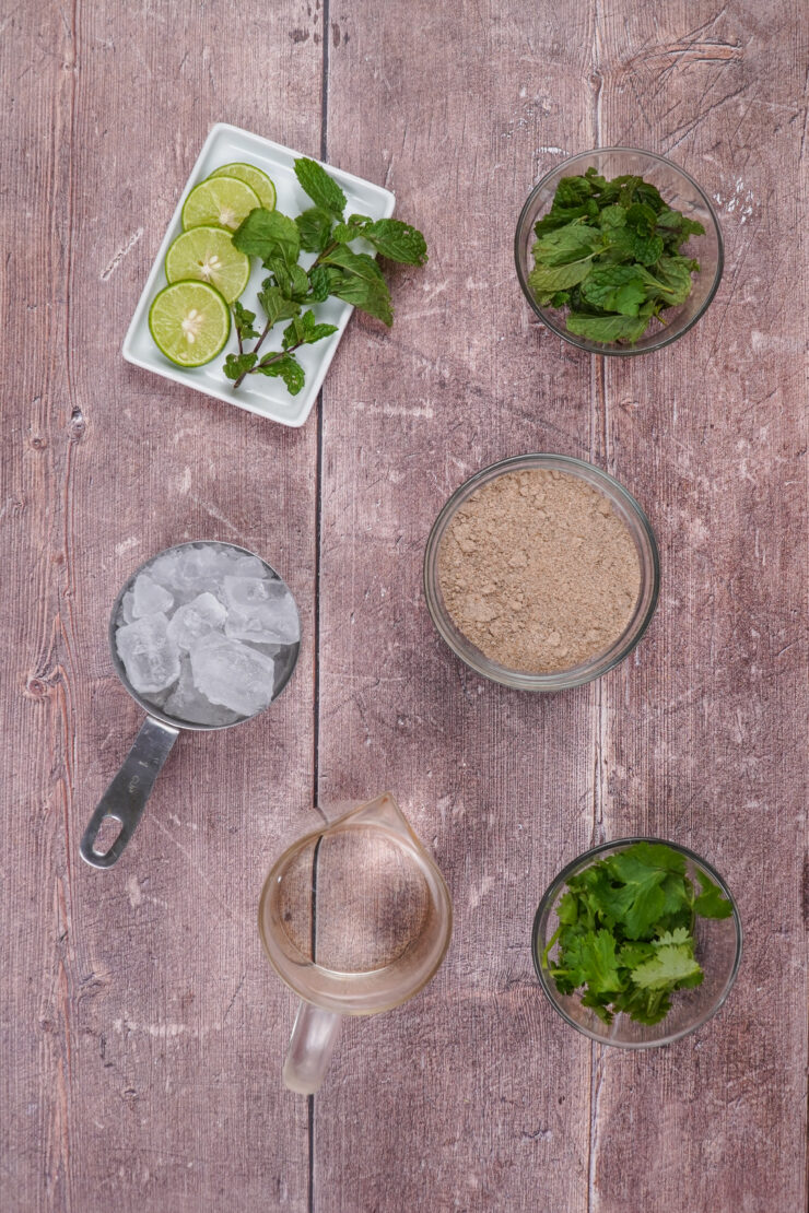 Overhead shot of bowls with the ingredients needed to make jaljeera including lime slices, mint sprigs, mint leaves, ice cubes, jeera powder, water, and cilantro leaves.